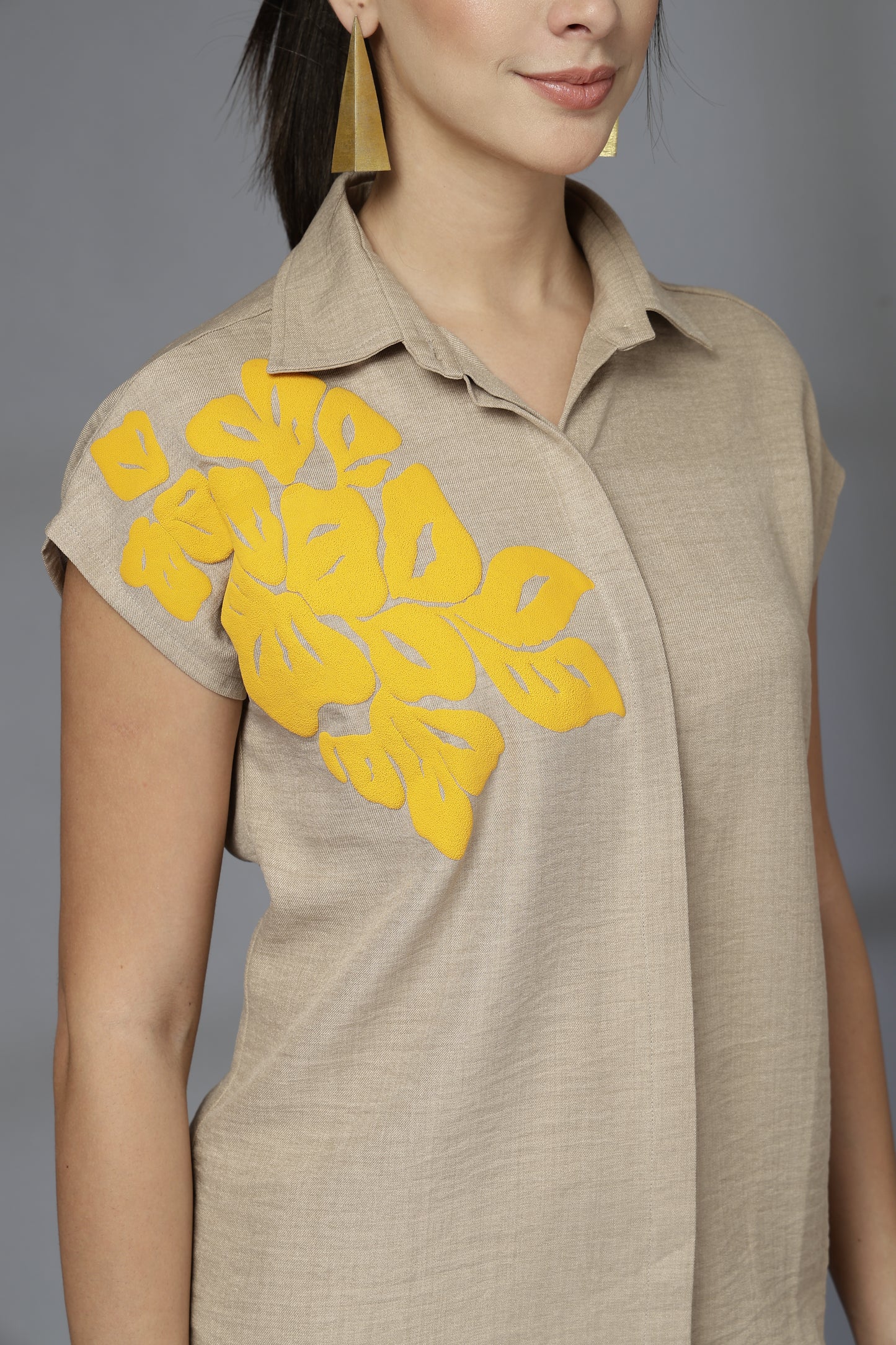 Woman wearing a beige sleeveless top with yellow floral appliqués on a gray background