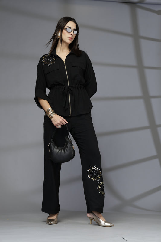 Woman in a black outfit with embellishments holding a black handbag against a gray background